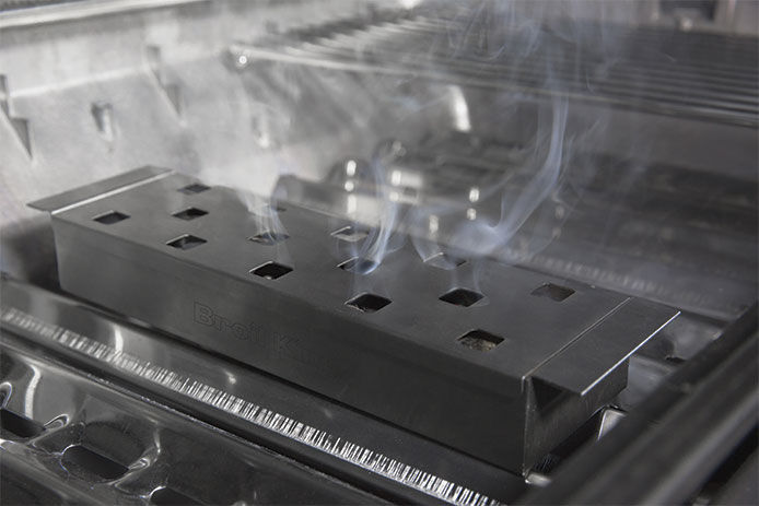 A silver smoking box smoking wood chips in under the grates of a gas grill