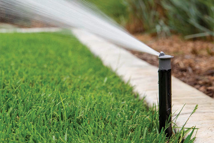 An inground sprinkler popped up out of the ground spraying water over a green lawn