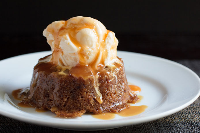 Close up image of sticky toffee pudding on top of a white plate isolated on a black background. The pudding is topped with vanilla ice cream and carmel syrup