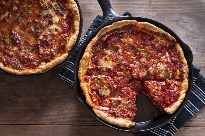 Two saucy deep-dish pizzas are shown in cast-iron skillets. The pizzas are resting on a wooden counter atop a black and grey striped towel.