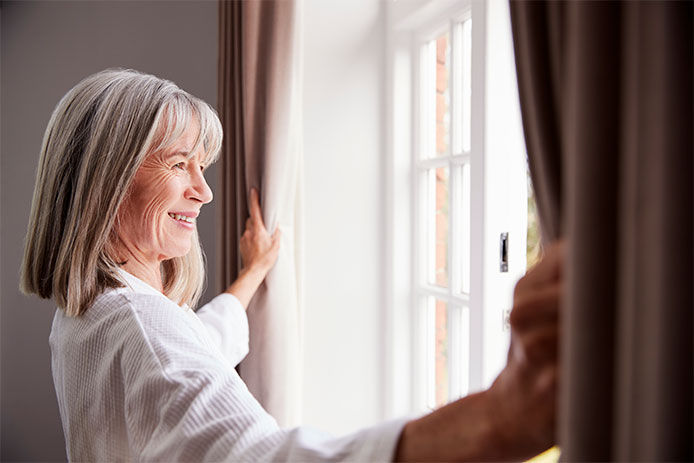 Senior Woman Opening Bedroom Curtains And Looking Out Of Window 
