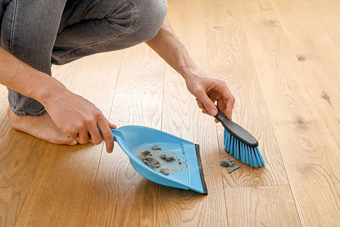 Person crouched using a handheld brush to sweep up dust bunnies into a small dust pan