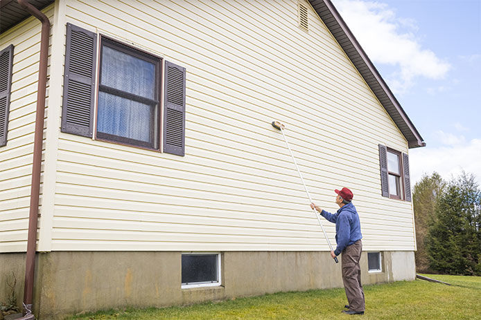 An elderly man using a telescoping brush to clean the vinyl siding on his home