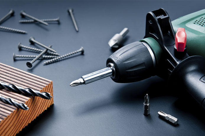 An electric drill, drill bits, screws, and a wooden board arranged together against a dark background. The electric drill is a corded model with a black handle and a metallic drill bit attached to the end. 