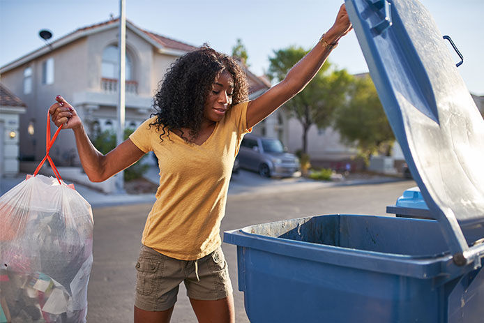 A woman wearing a tan shirt and brown cargo shorts lifts the blue lid of a trash can. She holds a trash bag in one arm, ready to deposit it in the bin. In the background, there is a stucco house, trees, and a car.