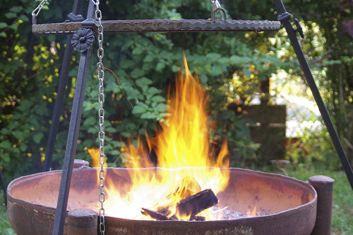 A fire pit is shown with a black cast iron tripod and chain attached to the top. The fire pit is burning in an open area with greenery and trees in the background.