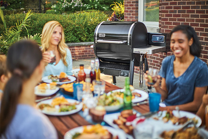 An outdoor party with young womena sitting around a table and a gas grill in the background 