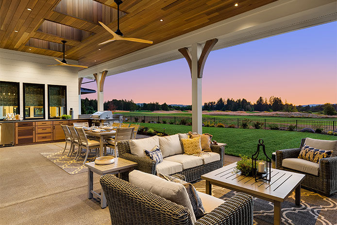 Luxury home exterior at sunset: Outdoor covered patio with kitchen, barbecue, dining table, and seating area, overlooking grass field and trees.