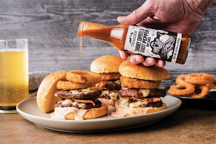 A close up image of Traeger Smoky Chipotle & Ghost Pepper sauce being poured onto a double cheeseburger with onion rings on top 