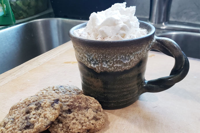 A brown mug with hot chocolate and whipped cream on top beside some cookies