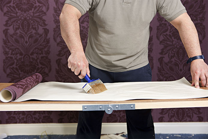 An older man using a 3" glue brush to apply wallpaper glue to the back of a strip of wallpaper 