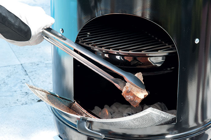A close up image of a pair of tongs being used to add chunks of wood to a Weber Smokey Mountain Cooker Smokers
