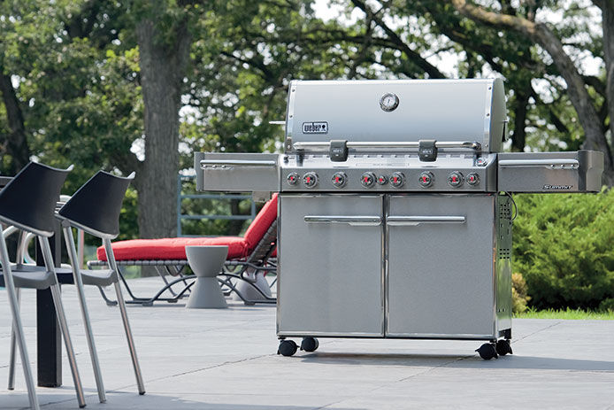A Weber stainless steel eight burner Summit gas grill sitting on a stone patio with patio furniture in the background