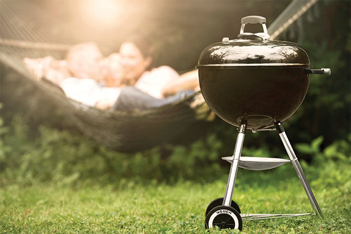 A black Weber Original Kettle Charcoal grill sitting in the grass ouf a backyard with a blurred out image of a family laying in a hammock in the background