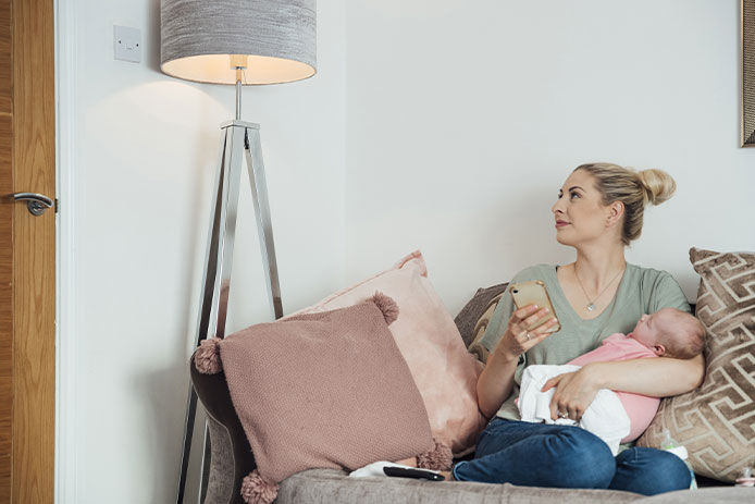 Woman sitting on the couch holding her baby in one arm while turning on her lamp with her phone in the other hand