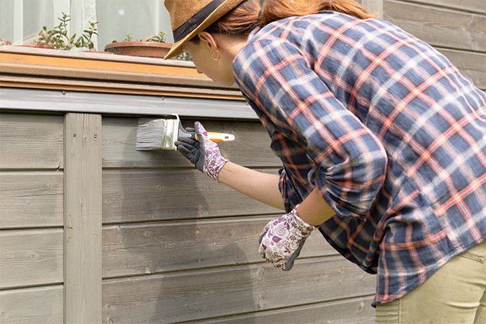 A woman wearing a flannel shirt and garden gloves painting the side of her home with a paint brush 