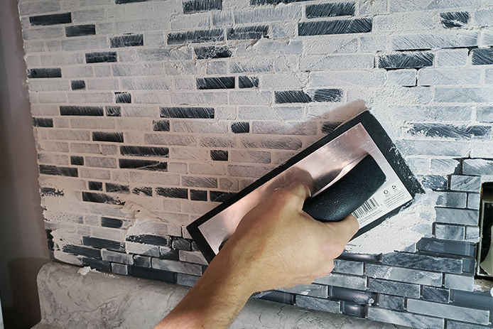 A close up of a man's hand applying grout over glass tile backsplash using a grout float