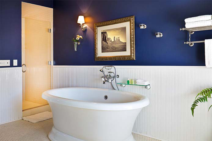 A modern contemporary classic bathroom design, furnished with a classic design, sepia toned picture on the wall, wainscoting, a freestanding pedestal bathtub and a modern shower stall, classic tile pattern on the floor. Photographed in horizontal format.