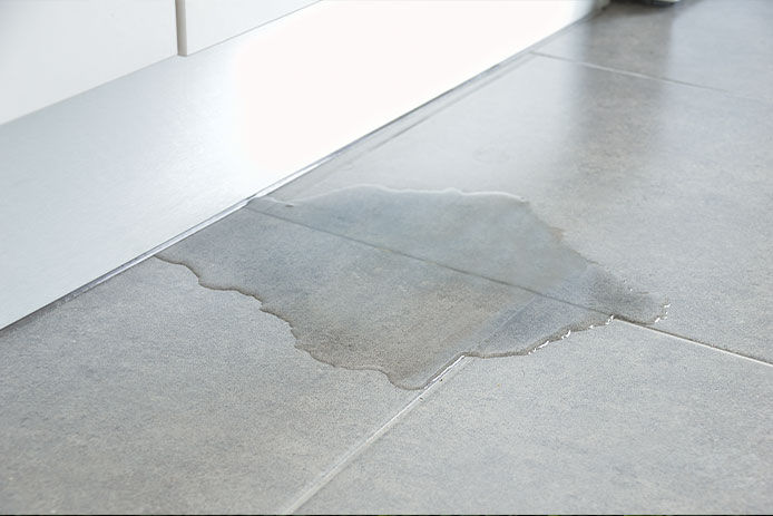 Close-up photo of flooded kitchen floor from water leak with damaged wooden cabinets and water puddles on tiles.