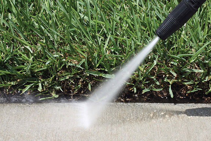 The black nozzle of a power washer sprays out a strong stream of water to wash a concrete sidewalk. The grass is shown in the background.