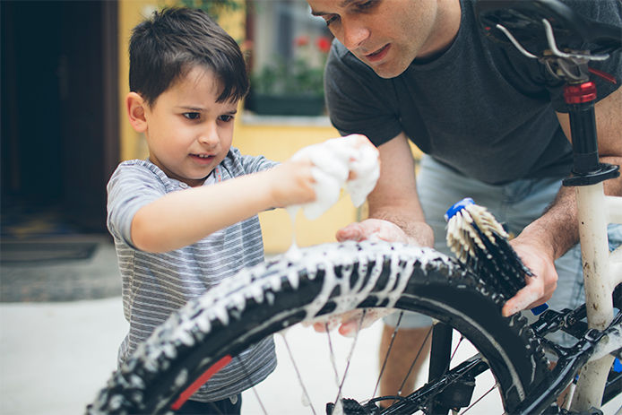 This image shows the cropped view of a father and son cleaning a bike tire. The focus of the image is on their hands, which are holding a damp soapy cloth above the black tire. 