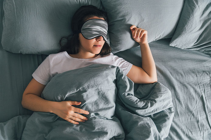 A middle aged woman laying in a bed with gray sheets and wearing a sleeping mask