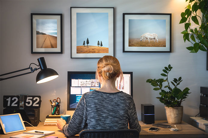 Woman sitting at her office desk with back facing the camera, desk lamp turned on and working on her laptop