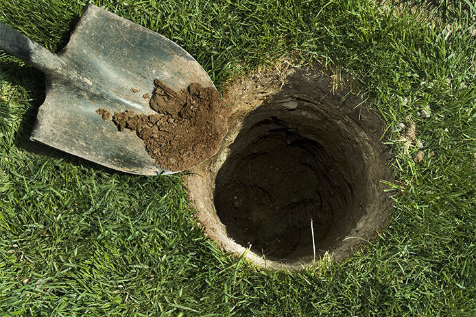 A grey metal shovel is shown next to a deep round hole that someone has dug in the middle of green grass.