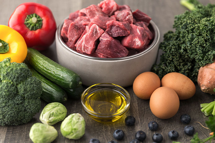 Raw dog food ingredients including, raw meat, brussel sprouts, eggs, blueberries, broccoli, peppers, oil, and cucumbers