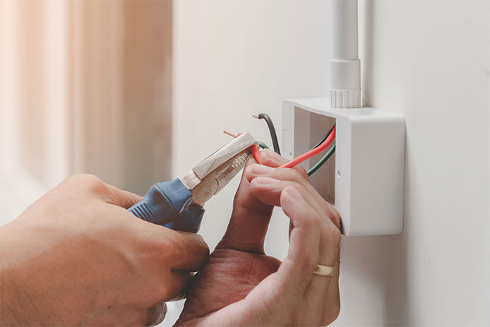 A person uses a set of wire strippers to remove the wire insulation coating on a red wire in a light socket box. 