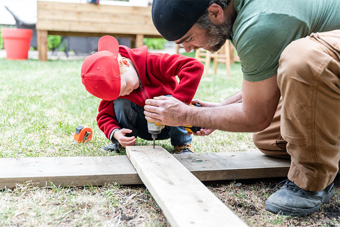 A father and son using a cordless power drill to drill screws into two boards of lumber to build a playgound set.