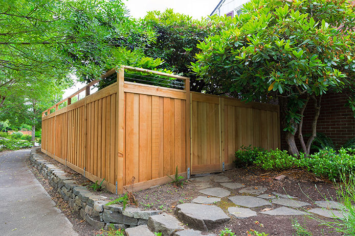 A rural neighborhood residential home with tall trees and a paver retaining wall that surrounds a tall wood slated fence