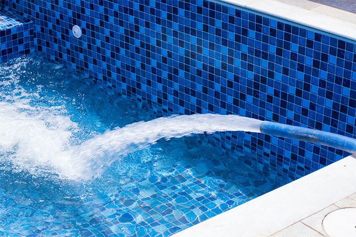 A blue tiled swimming pool is being filled with clear blue water from a blue garden hose. The pool is surrounded by a concrete deck.