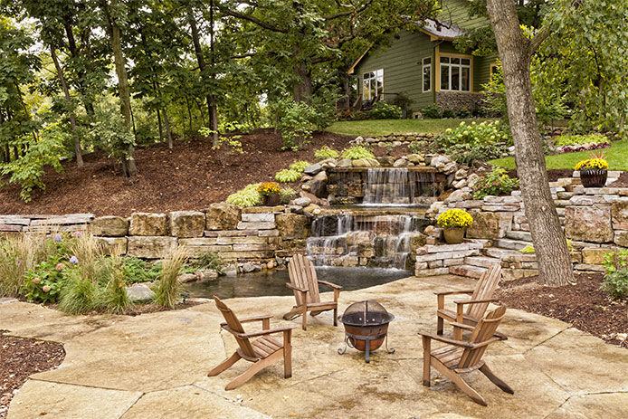 Beautiful landscaping with waterfall, koi pond, and stone patio with wooded surroundings.