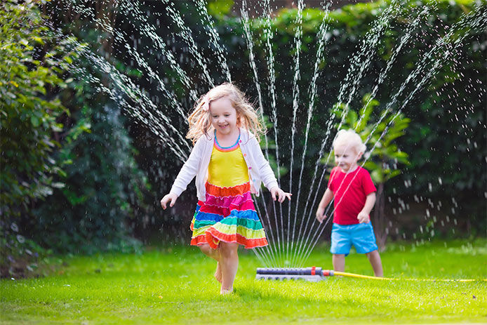 Child playing with garden sprinkler. Preschooler kid running and jumping. Summer outdoor water fun in the backyard. Children play with hose watering flowers. Kids run and splash on hot sunny day.