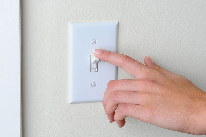 A hand is pressing a white light switch down. The switch is mounted on a cream-colored wall with white trim.