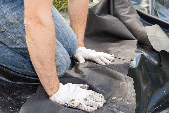 close-up of a man's hands wearing white gloves as he folds over a pond liner on the ground. The focus is on the liner, which is visible in the foreground. The pond liner is being folded neatly, indicating the man is taking great care in the installation of the pond. The man is wearing blue jeans, which can be seen in the background. This image is perfect for those interested in DIY pond installation or backyard landscaping.