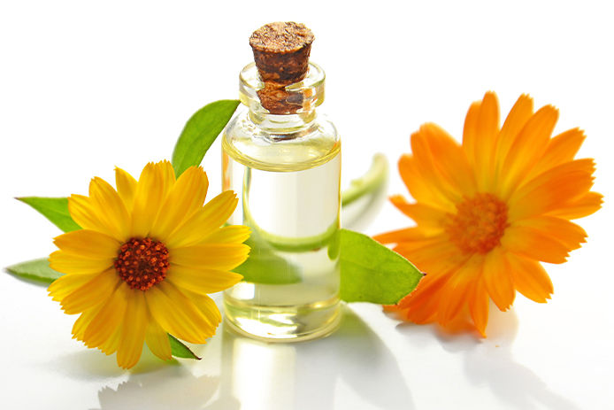 Homemade essiental oil glass bottle with two colorful flowers surrounding it. The glass bottle has a cork cap and is filled with clear liquid.