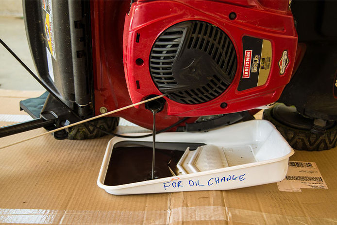 Lawn mower on its side draining oil into a pan with cardboard are the ground