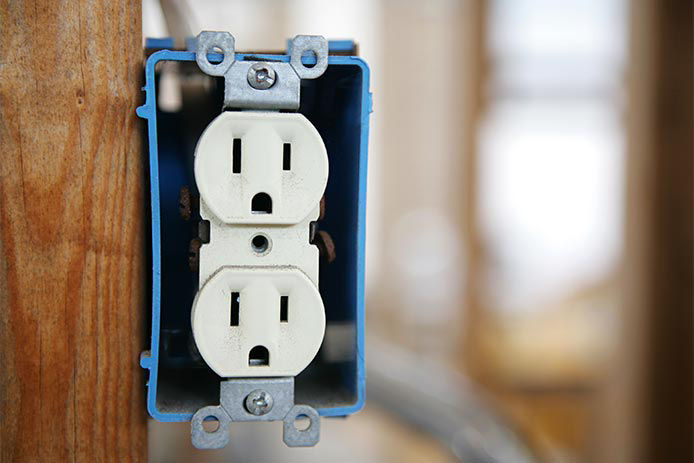 A blue box contains an electrical outlet mounted on the wooden frame of a garage. The outlet is white and grounded without a plate cover.