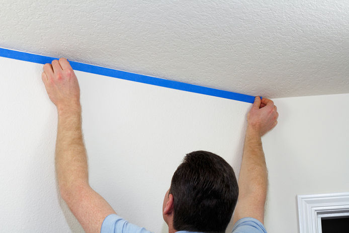 A man applying blue painters tape to the top of a wall underneith the ceiling 