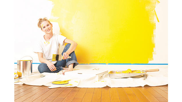Woman sitting on ground with back towards painted wall and paint brush in hand