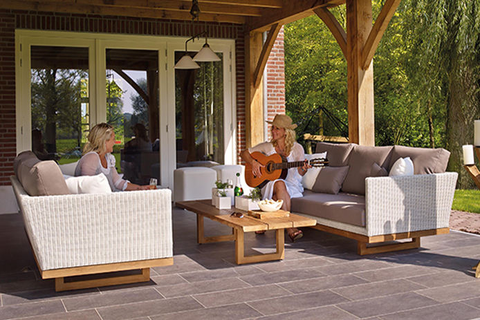 Two women sitting outside on beige and white patio furniture with wooden frames, playing the guitar and drinking champaign while laughing and having a great time.