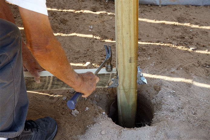 A person in a white shirt is placing a wooden post into the dirt. They’re using a hammer to tap the post into place with metal fittings.