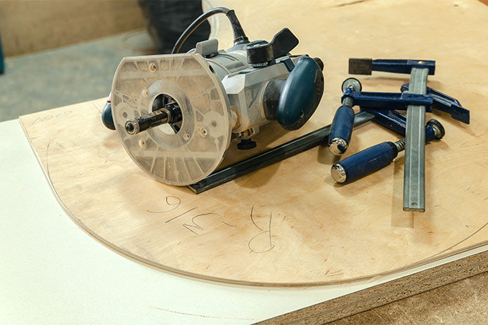 A silver-colored wood router rests atop a curved piece of light-colored wood. Several woodworking tools are piled next to the router. 