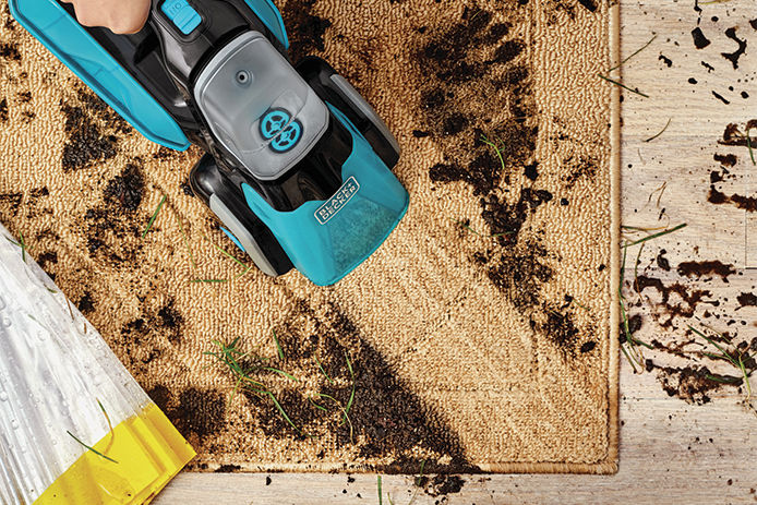 A teal handheld Black & Decker carpet cleaning cleaning up mud, dirt, and grass off of a beige rug.