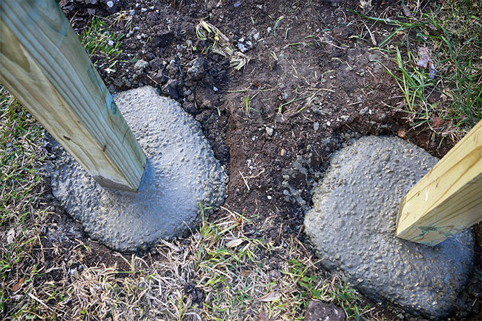 Two wooden posts are shown, nestled into concrete bases. The concrete is set and sturdy, and the posts are buried in the dirt and grassy ground.