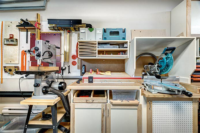 A garage woodworking station is filled with white and wooden cabinetry, colorful tools, and shelving. The workstation features a workbench with saws, drills, and other tools.