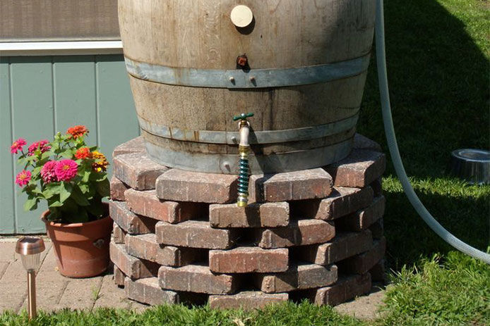 Wooden rain barrel sitting on a stack of brown and red bricks with a hose extention on the bottom spigot 