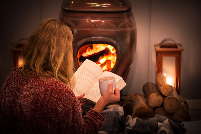 Woman sitting by wood stove reading her book with coffee in hand